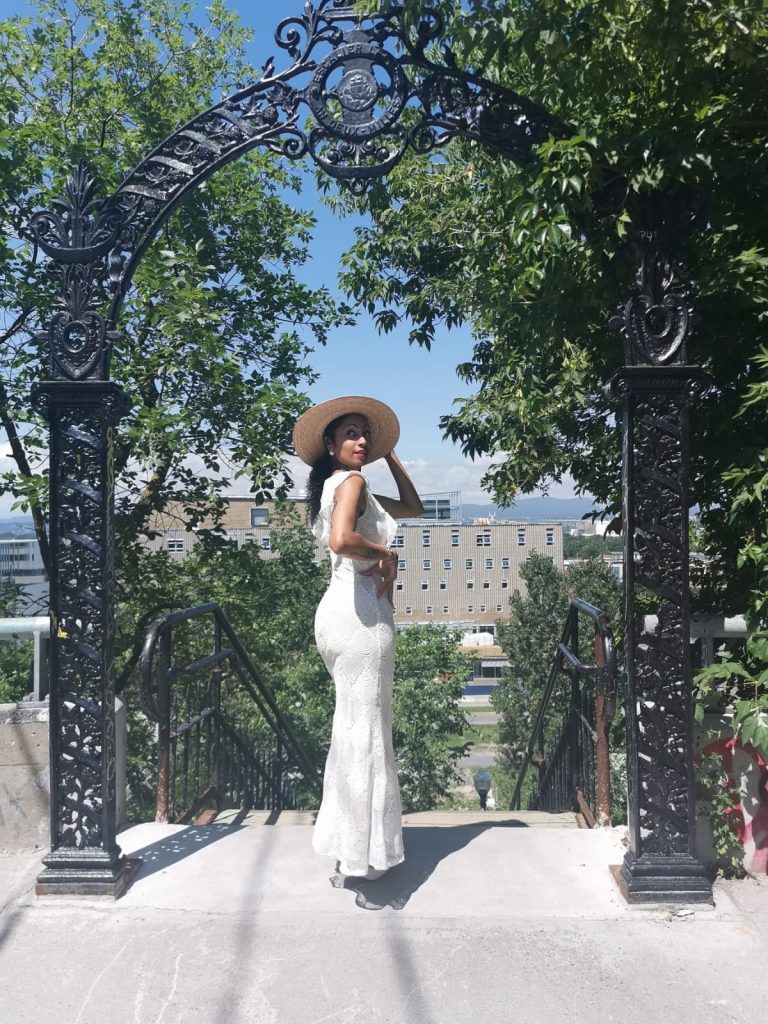 A stylish woman in a white dress and hat posing by an ornate gate in Quebec city