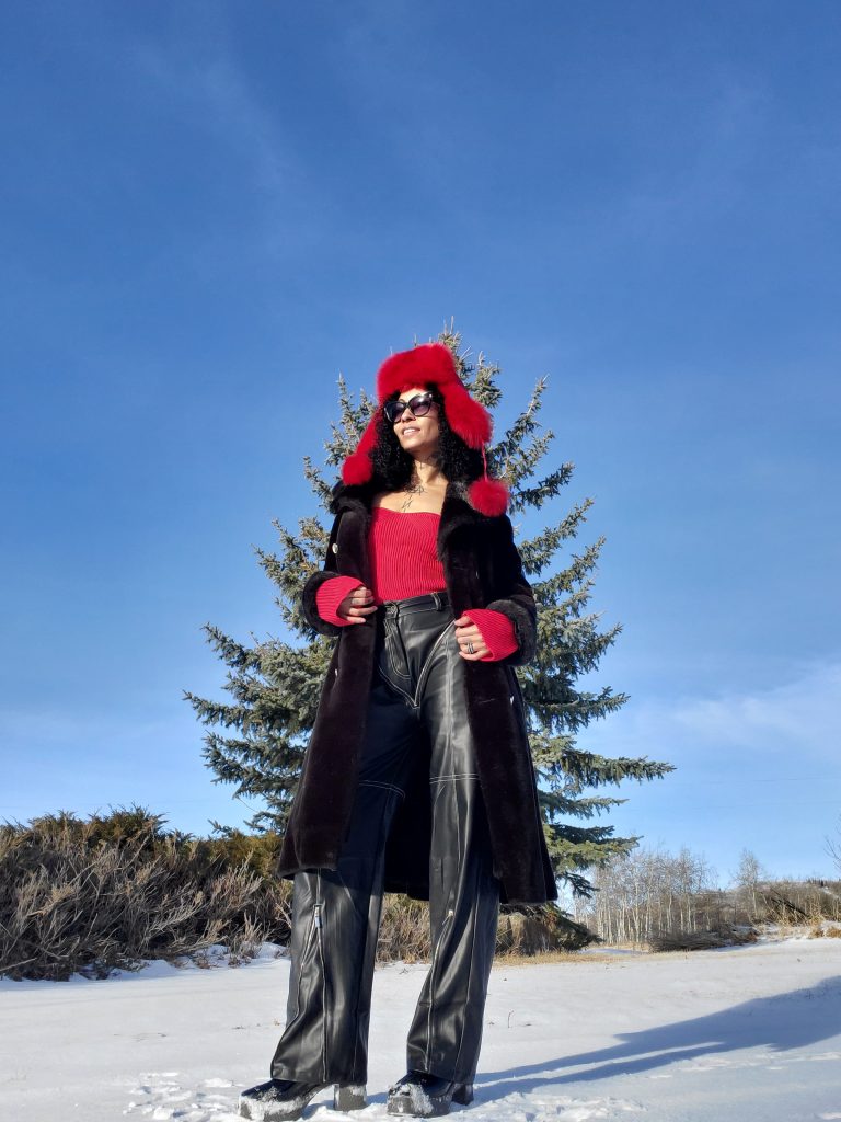 Elegant lady in red sweater and black pants sits gracefully on snow, accessorized with a stunning red fur Russian winter hat.
