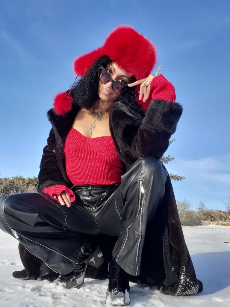 Woman in red sweater and black pants sits on snow, wearing matching red fur Russian winter hat for a luxurious winter look
