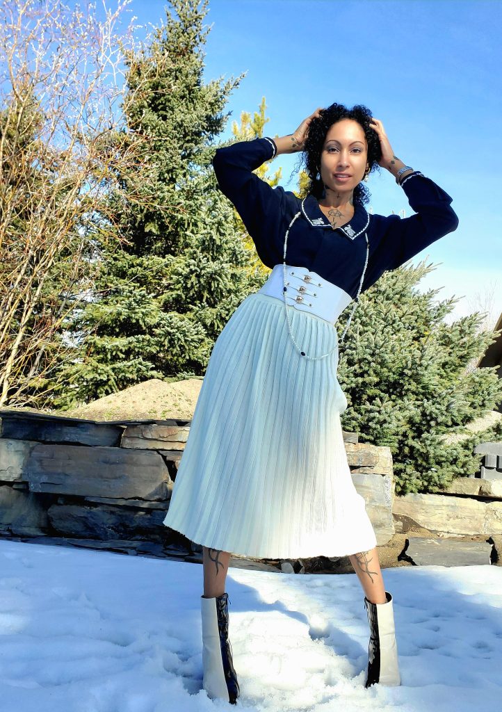 Elegantly clad in a 1970s vintage pleated skirt, a woman exudes timeless charm. Her embroidered blouse adds a touch of sophistication, while the PVC ankle boots lend a modern twist. A picturesque snowy backdrop completes this fashionable ensemble.