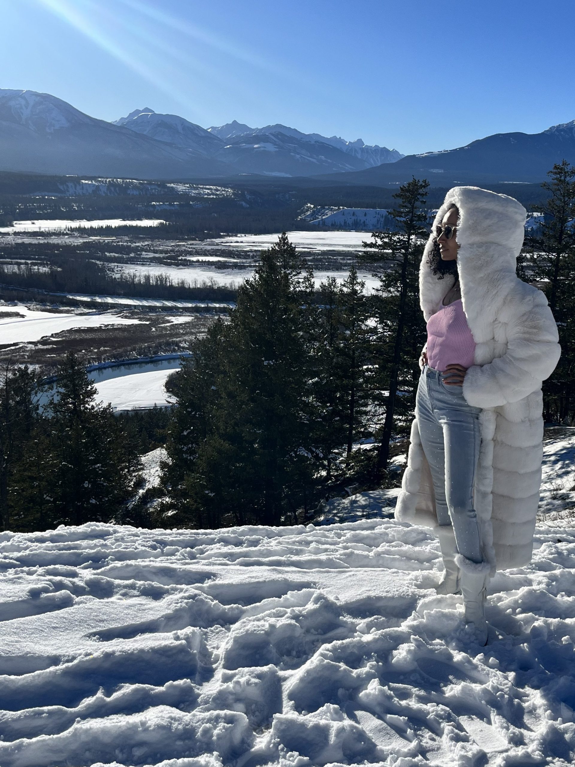 Fashionable woman in pink sweater and white fur coat poses on snowy mountain.