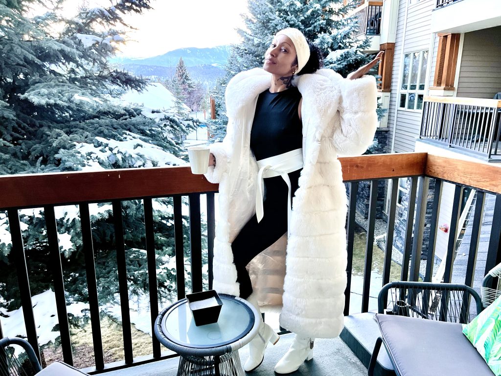 Stylish lady in white fur coat on balcony, dressed in black bodysuit and winter accessories.