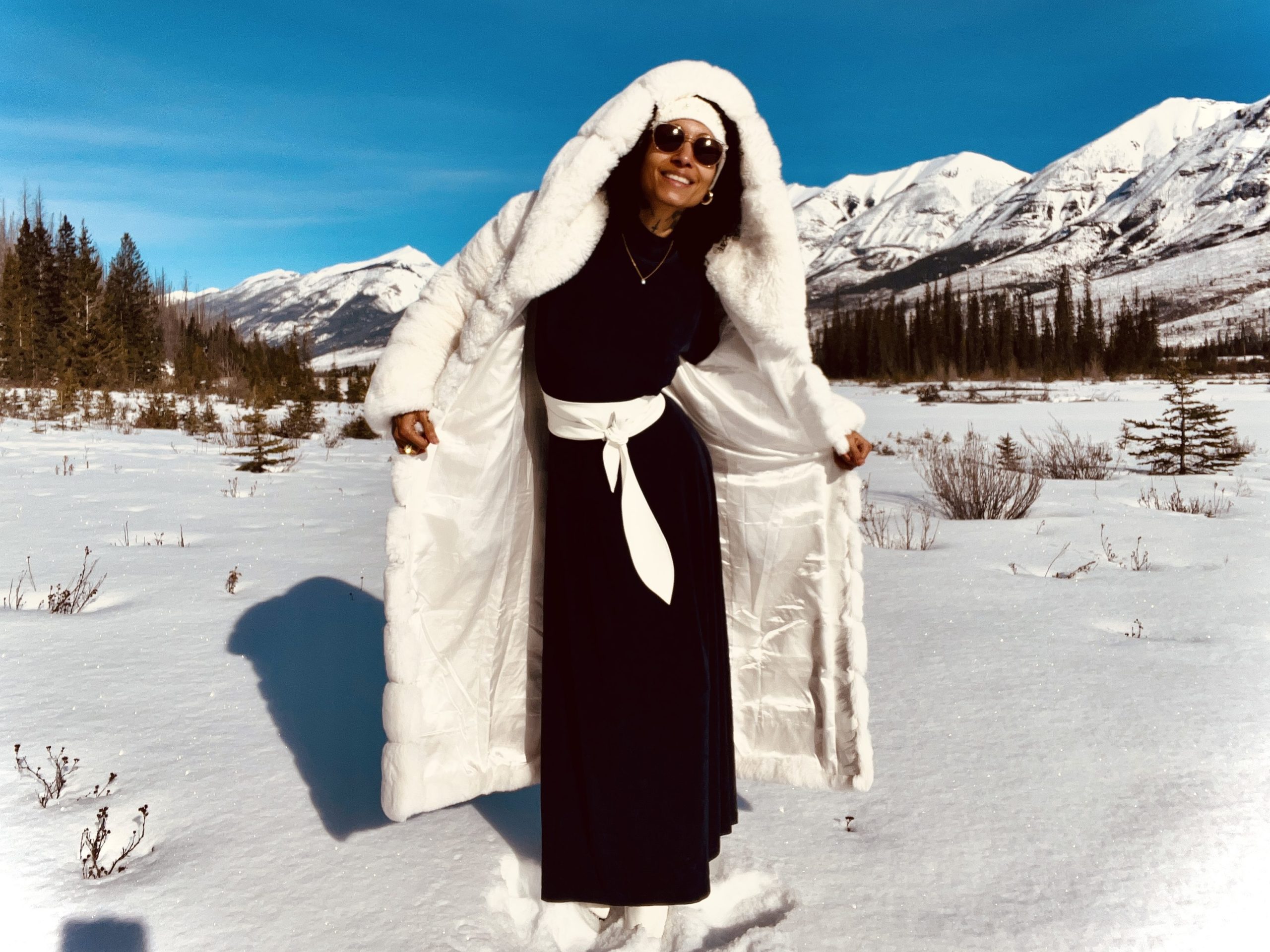 Elegant woman in white fur coat stands gracefully in snow, wearing dark blue vintage gown and white winter jacket.