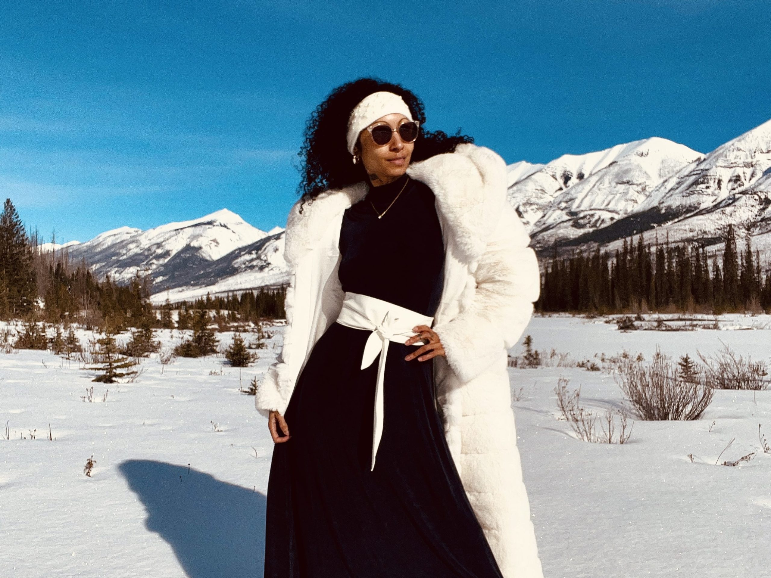 Woman exudes sophistication in white fur coat, standing in snow with dark blue vintage gown and white winter jacket.