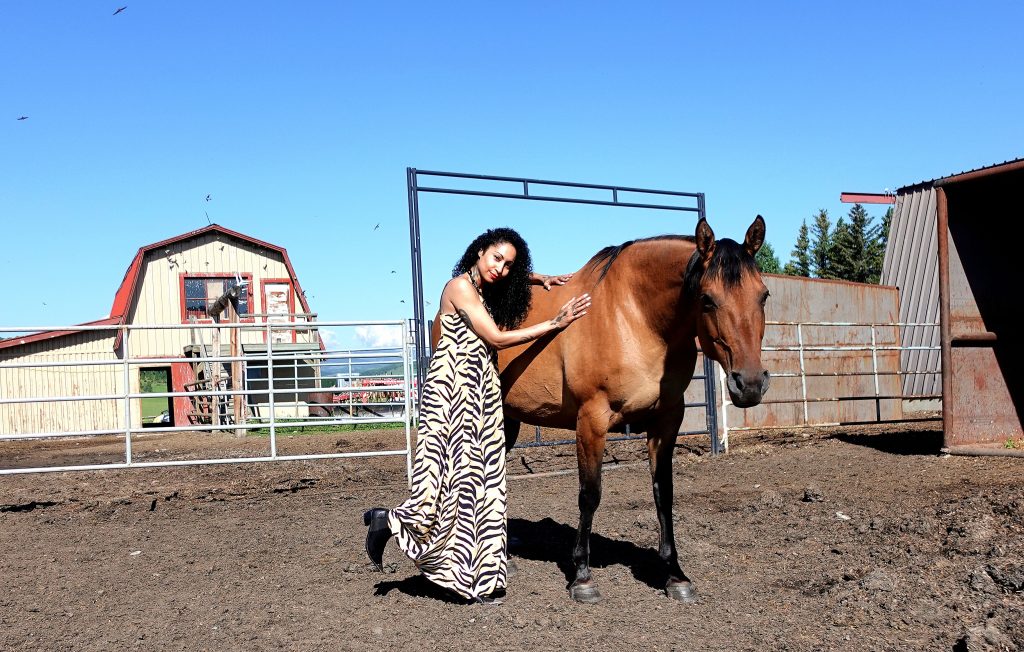 beautiful tattooed black model wears a tiger print dress, black steel tipped cowboy boots while petting a horse on a ranch